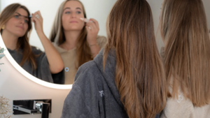 Deux jeunes femmes complices en poncho se maquillent dans la salle de bain, devant un grand miroir