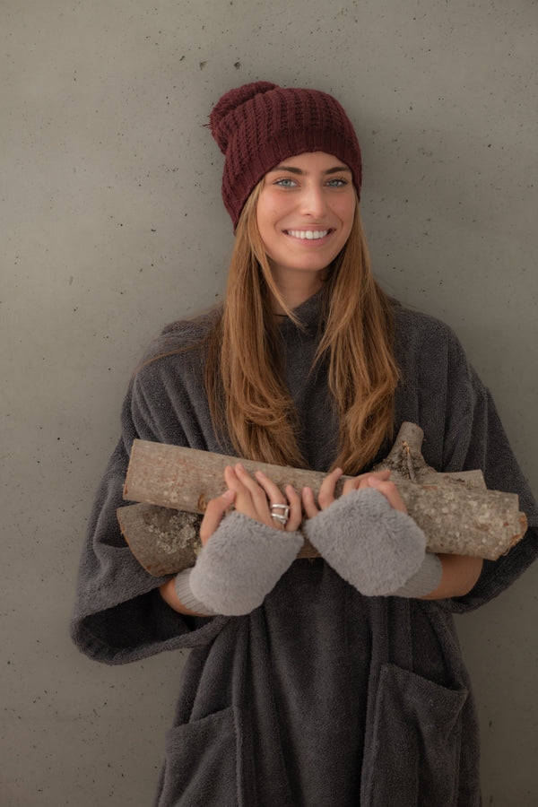Une jeune femme souriante en poncho bio graphite avec buches de bois dans les bras, portant un bonnet et des mitaines - Atelier Dune