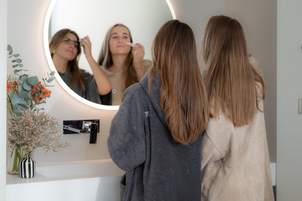 Deux jeunes filles se maquillent dans leur salle de bain devant un miroir et portent un poncho graphite et sable bio - Atelier Dune