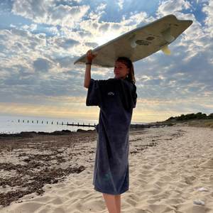 Jeune fille sur la plage en poncho de bain graphite portant une planche de surf sur la tête - Atelier Dune