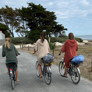 Trois jeunes filles de dos à vélo en ponchos de bain sauge sable et terracotta - Atelier Dune