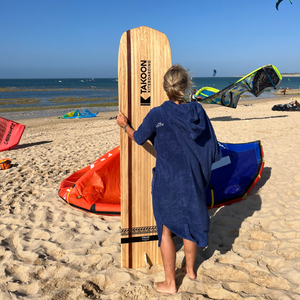 femme de dos sur la plage en poncho de bain outremer portant une planche de surf devant elle - Atelier Dune