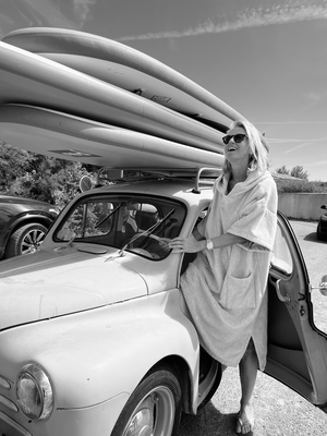 Fondatrice de la marque qui sort d'une voiture avec planches de surf sur le toit en souriant en poncho - Atelier Dune