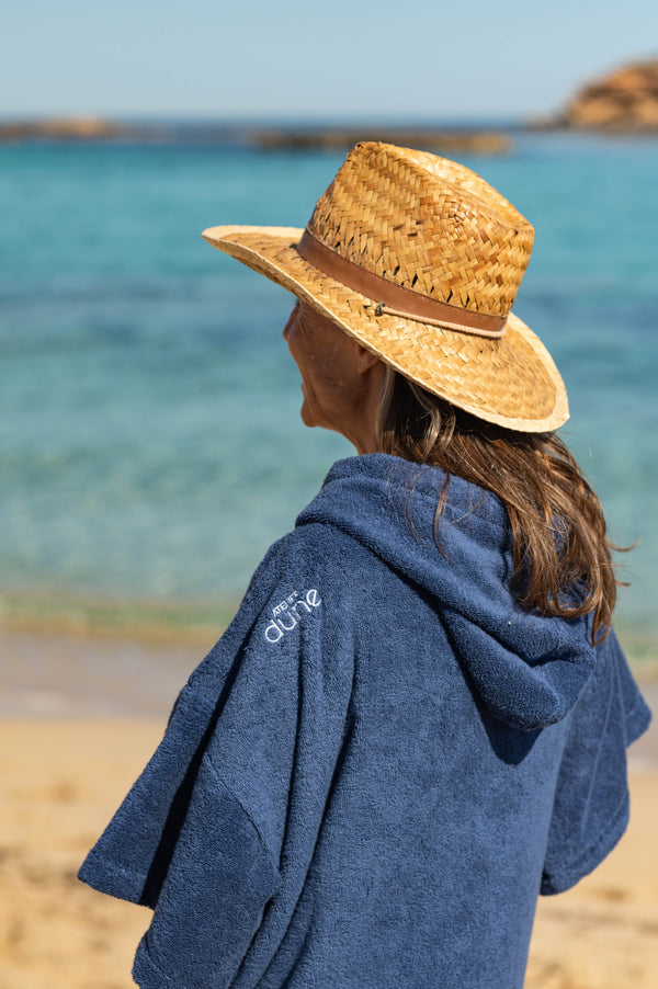 Femme de dos en bord de mer avec poncho outremer et chapeau de paille - Atelier Dune