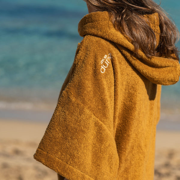 Jeune femme portant le poncho bio coloris caramel de dos, au bord de la mer - Atelier Dune