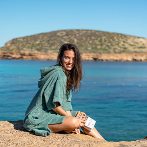 jeune fille souriante qui porte poncho de bain moelleux en coton biologique céladon assise sur un rocher devant la mer - Atelier Dune