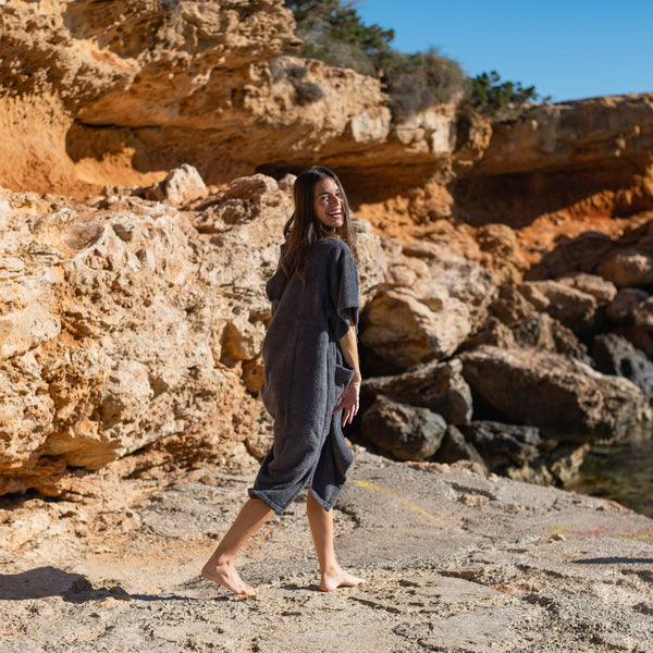 Jeune fille brune souriante marche dans un décor de calanque à Ibiza en poncho bio graphite - Atelier Dune