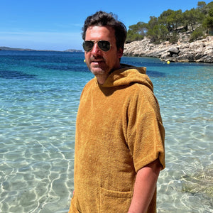 Homme souriant, portant le poncho bio coloris caramel les pieds dans la mer à Ibiza - Atelier Dune