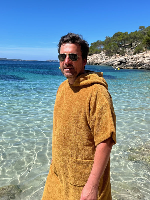 Homme souriant, portant le poncho bio coloris caramel les pieds dans la mer à Ibiza - Atelier Dune