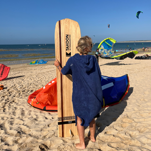 femme de dos sur la plage portant poncho de bain outremer et tenant plance de surf en bois - atelier dune