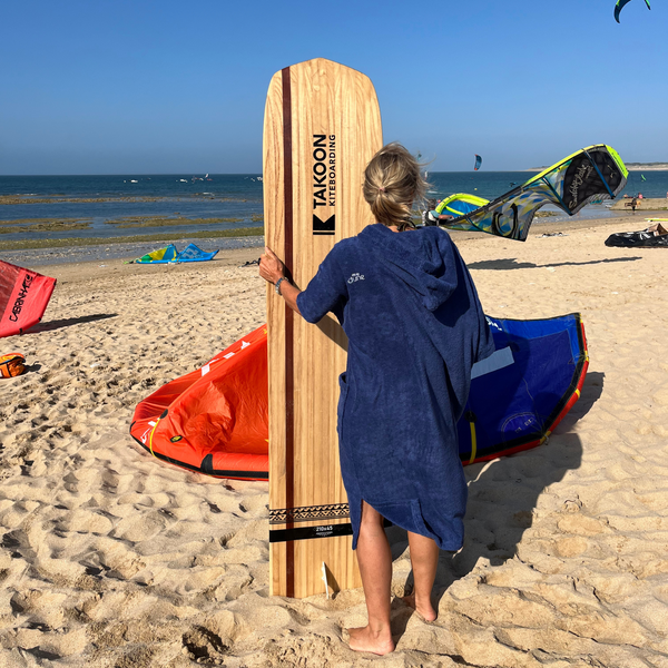 Femme de dos sur la plage en poncho de bain outremer avec une planche de surf - Atelier Dune
